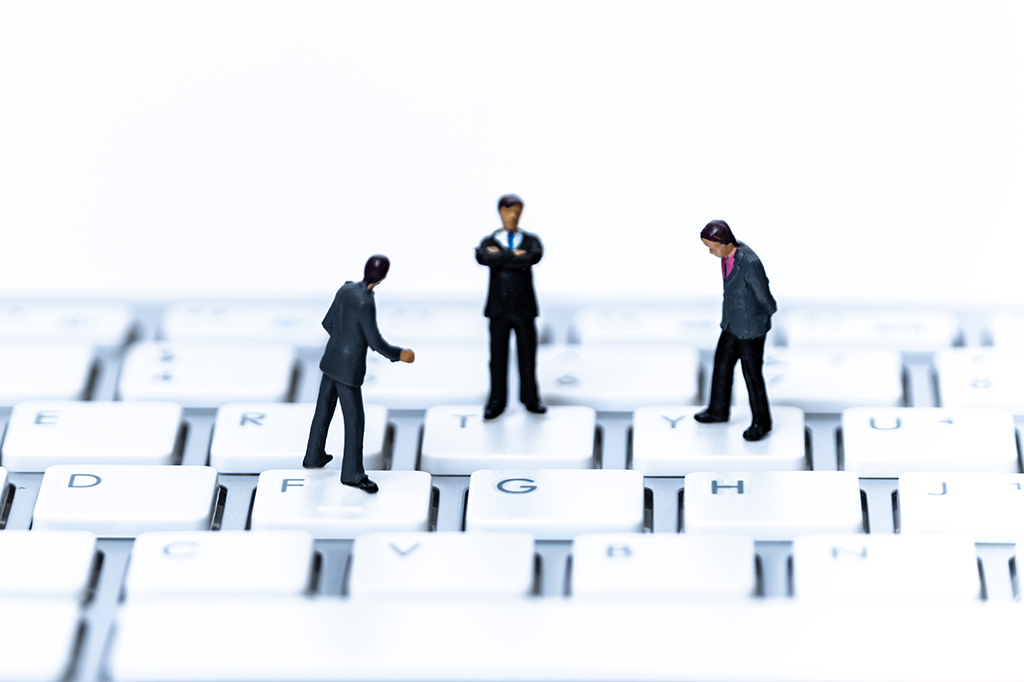 Três bonecos de miniatura, representando homens de terno, estão sobre um teclado de computador branco. Um está de costas, outro de braços cruzados no centro, e o terceiro de perfil, olhando para baixo. O fundo é branco, e as teclas têm letras como F, G, H, T, Y visíveis