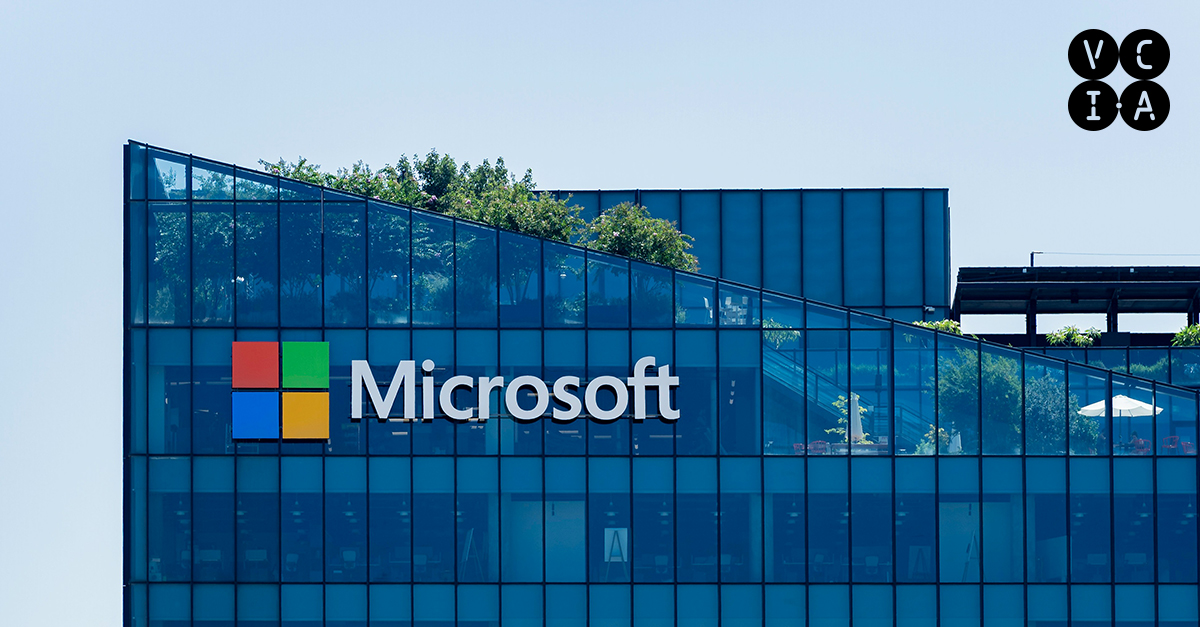 Fachada de um prédio moderno com janelas azuis espelhadas, exibindo o logotipo da Microsoft em branco e colorido. Há vegetação nos terraços e um guarda-sol branco visível